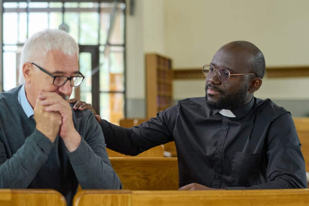 Young black man in shirt with clerical collar keeping hand on shoulder of senior male parishioner while comforting him and praying. Young black man in shirt with clerical collar keeping hand on shoulder of senior male parishioner while comforting him and praying.