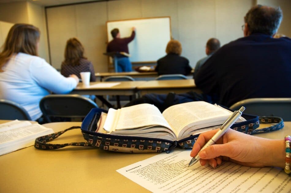 a bible in a custom fabric cover sits open on a desk with a teacher writing on a whiteboard in the background. a students hand holding a pencil writing on white paper is in the foreground on the table with the bible a bible in a custom fabric cover sits open on a desk with a teacher writing on a whiteboard in the background. a students hand holding a pencil writing on white paper is in the foreground on the table with the bible