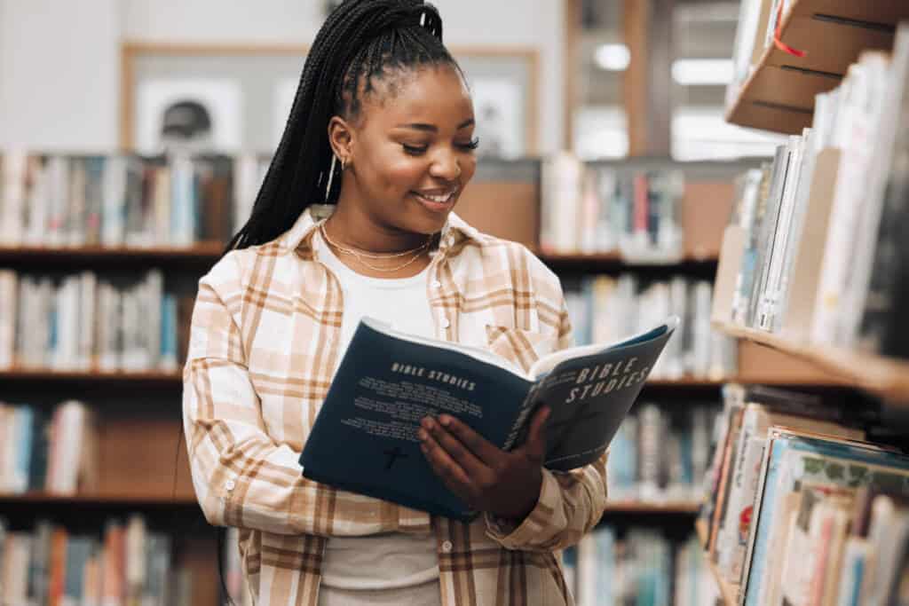 a woman with dark braided hair and a plaid shirt holds a book called "bible studies" open in a library