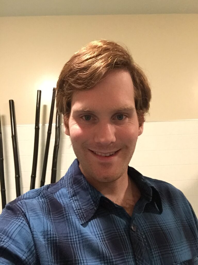 A man with brown hair wearing a blue plaid shirt smiles at the camera indoors.