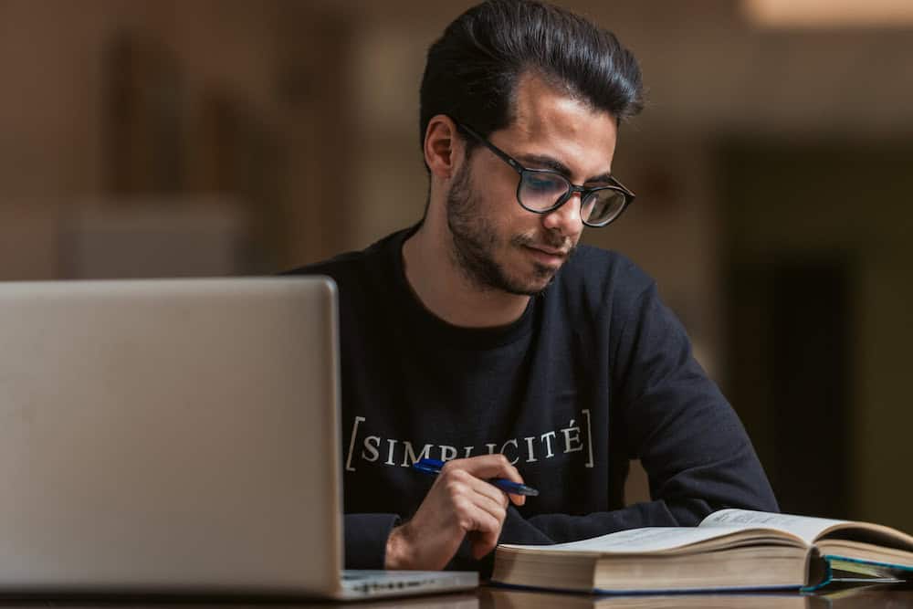 Man wearing glasses studies at a laptop with an open book beside him.