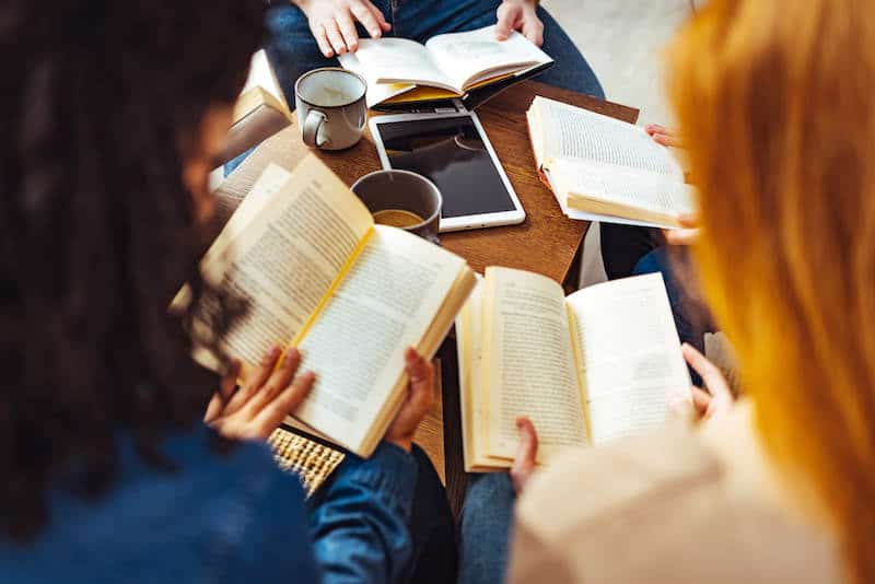 Three people sit at a wooden table reading open books with a smartphone and mugs nearby.