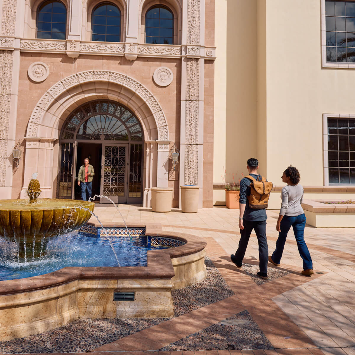 Two people walk past a fountain in a courtyard with arched doorways and terracotta buildings.