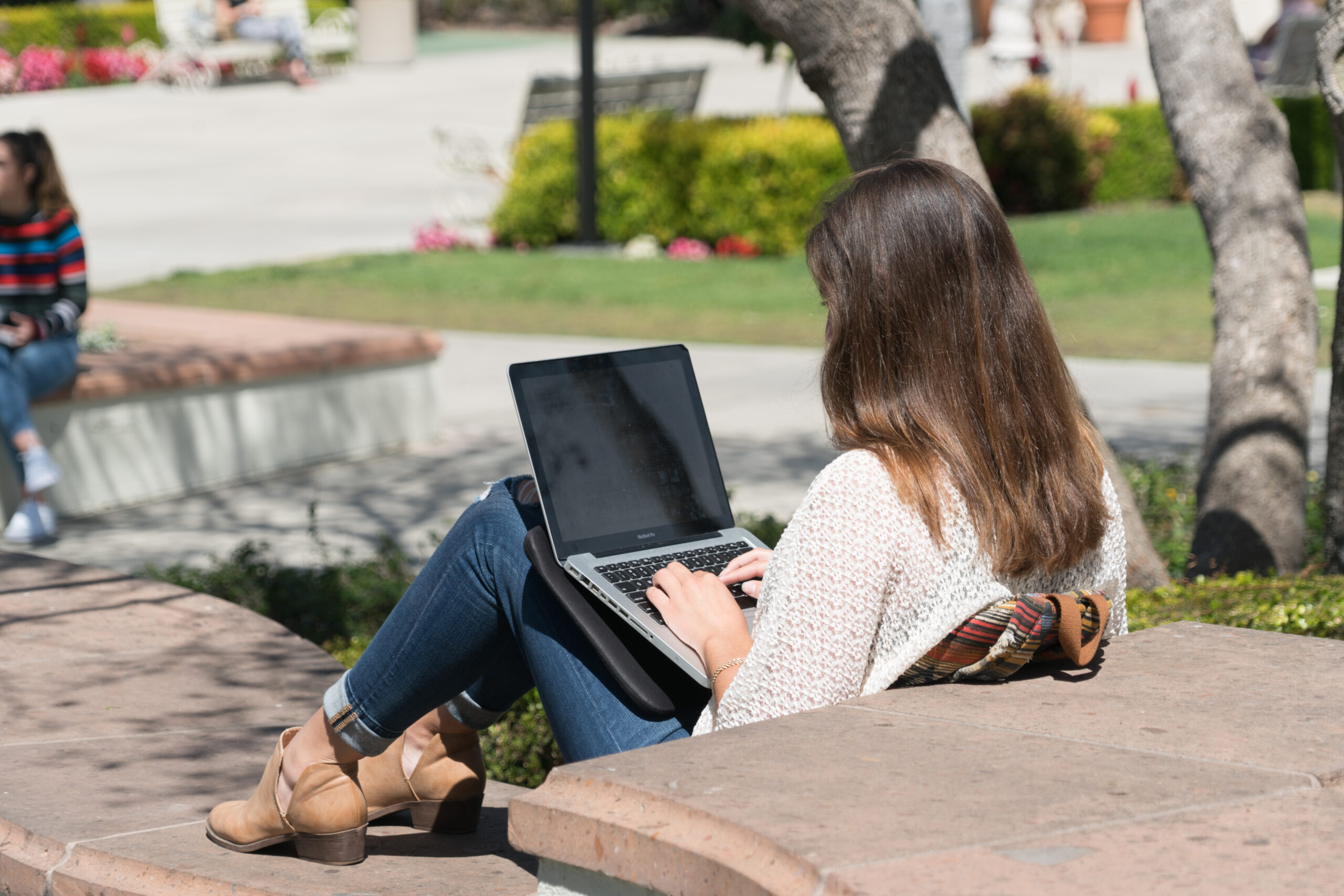 Woman sitting outdoors on a bench using a laptop in a park setting.