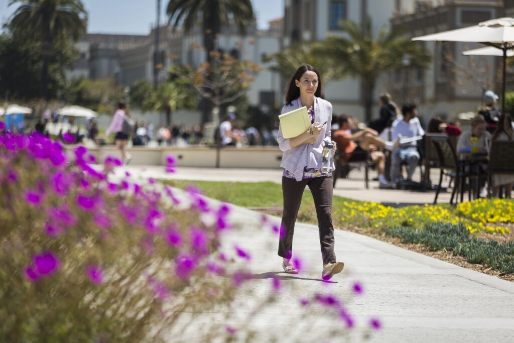 Woman jogging through a sunny urban plaza with purple flowers and palm trees.