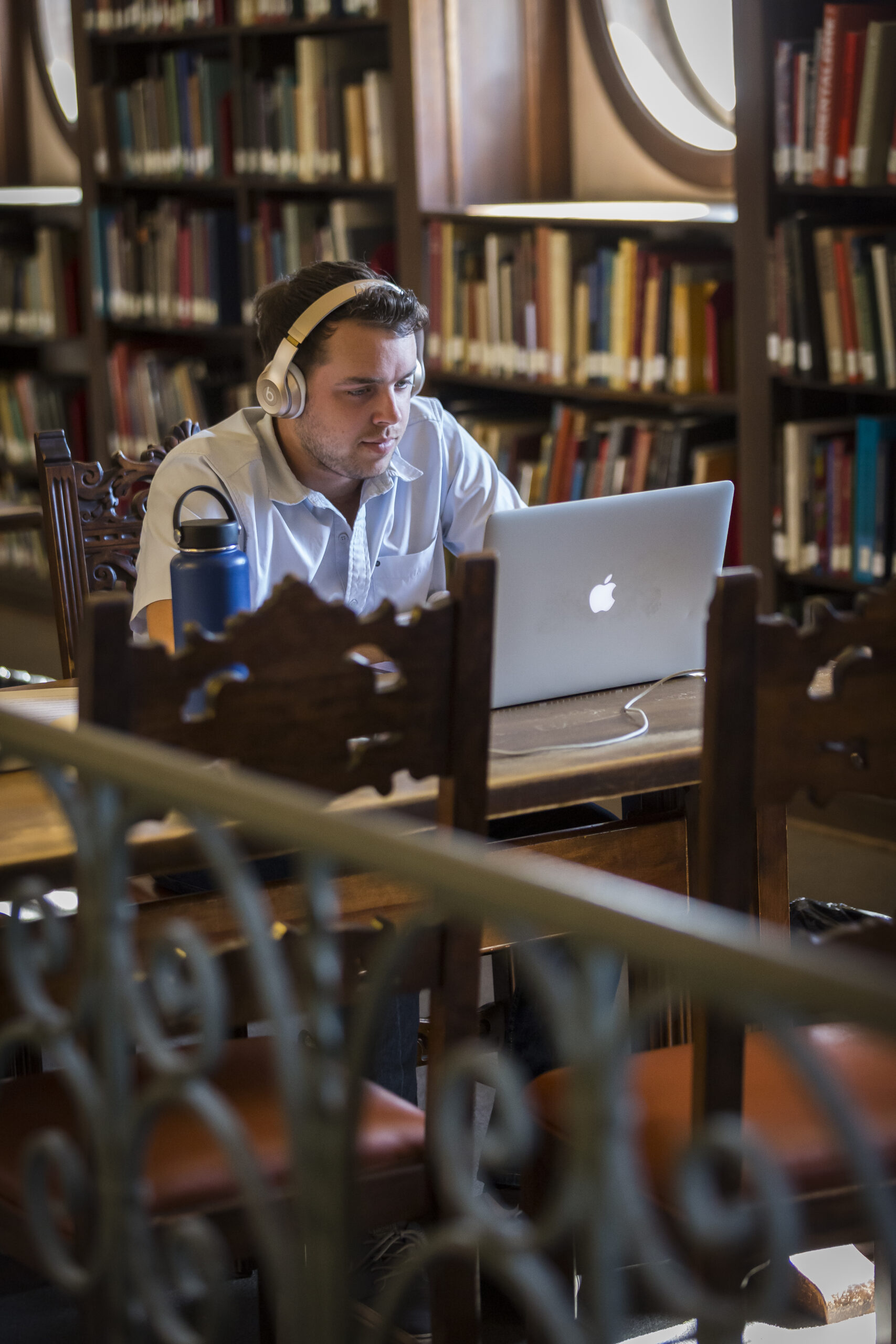 Man wearing headphones works on a laptop in a library with bookshelves in the background.