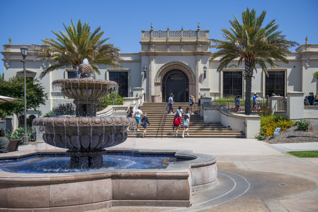 A grand white building with classical architecture, flanked by palm trees, with visitors on its.