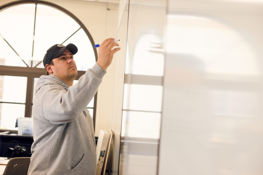 Man in a gray hoodie and cap adjusts a blue object on a white wall indoors.