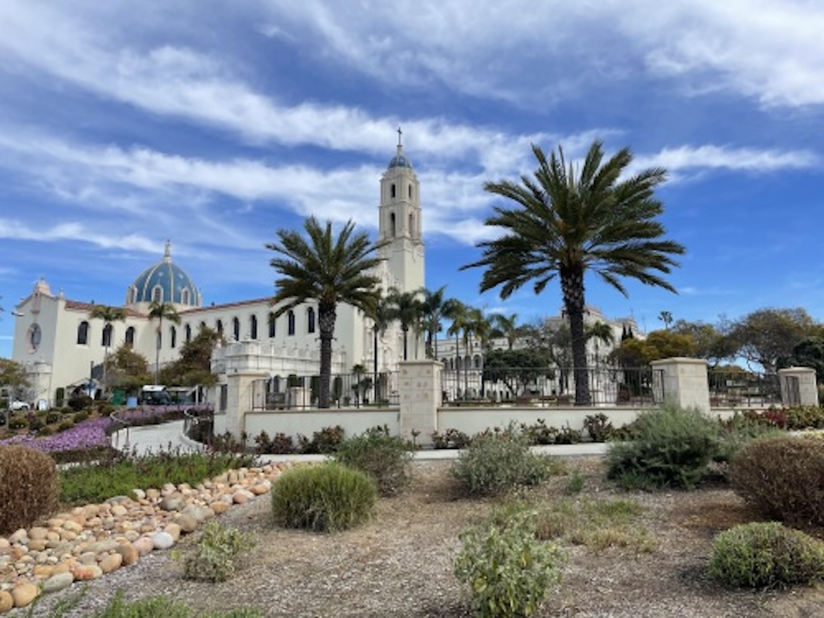 White Spanish colonial church with tall bell tower surrounded by palm trees and desert landscaping.