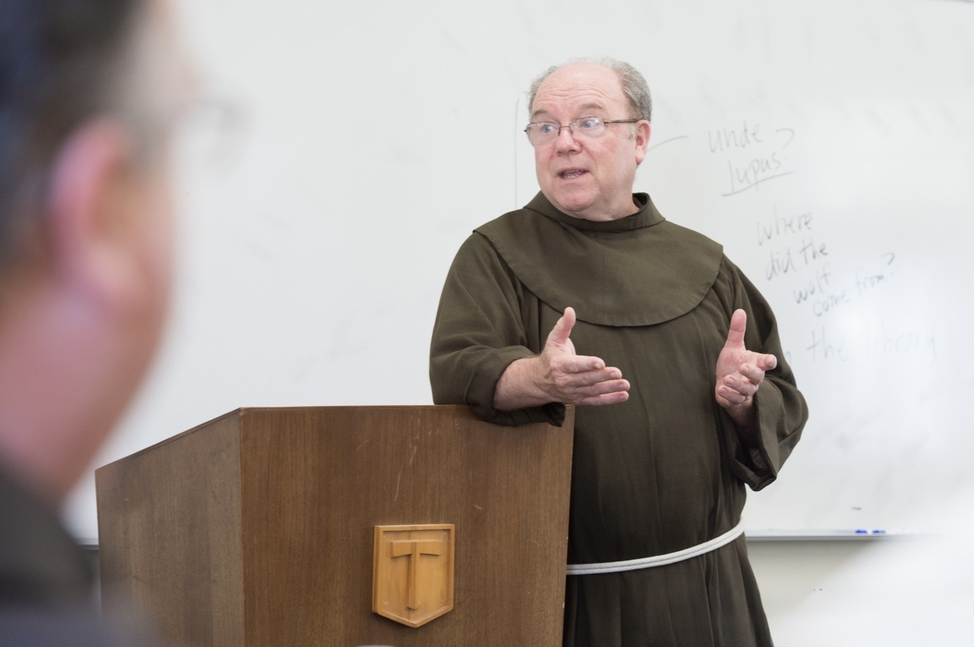 A man in a brown robe speaks at a wooden podium with a cross emblem.