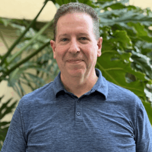 A man wearing a blue polo shirt stands in front of green foliage.