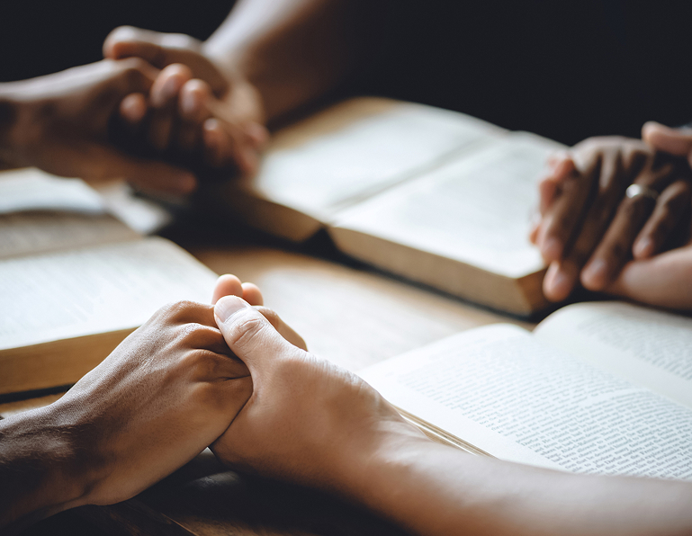 Multiple hands rest on open books during a group study or prayer session.