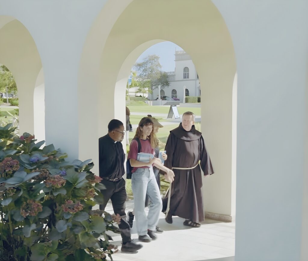 Three people walk through a white arched corridor toward a white building with a bell tower.