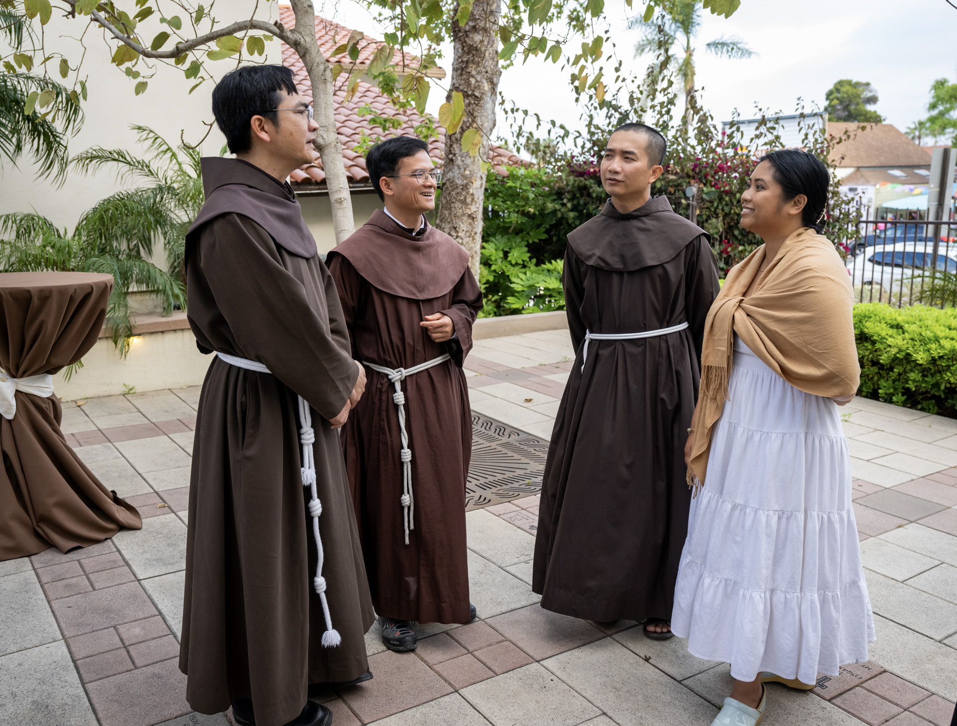 Four people stand in a courtyard, three wearing brown robes and one wearing a white skirt and tan.