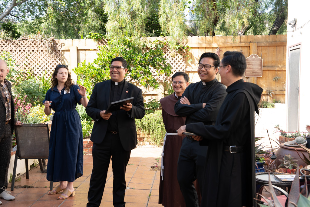 A group of clergy and religious figures gather in a garden courtyard during an outdoor ceremony.