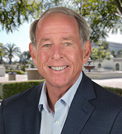 Smiling man with gray hair wearing a dark blazer and light blue shirt outdoors.