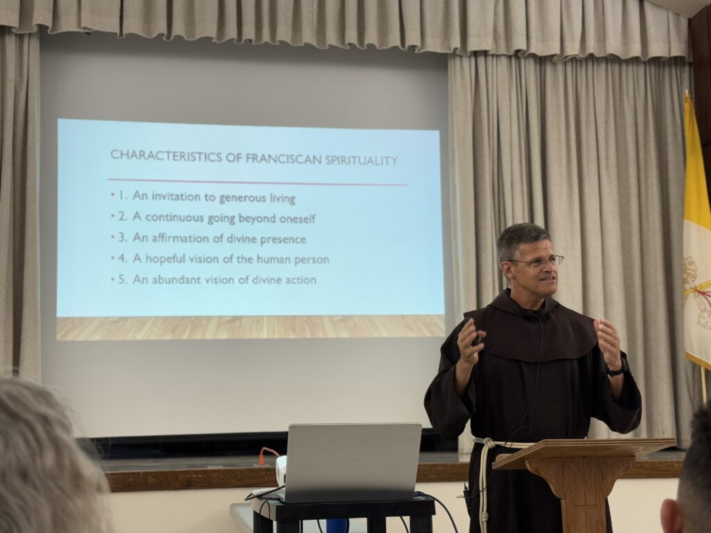 A man in brown robes presents at a podium with a projected slide listing characteristics.