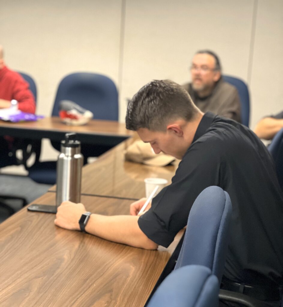 A young man in a black shirt sits at a conference table during a meeting.