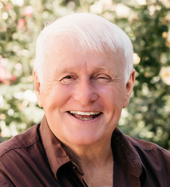 Smiling older man with white hair wearing a dark jacket outdoors.
