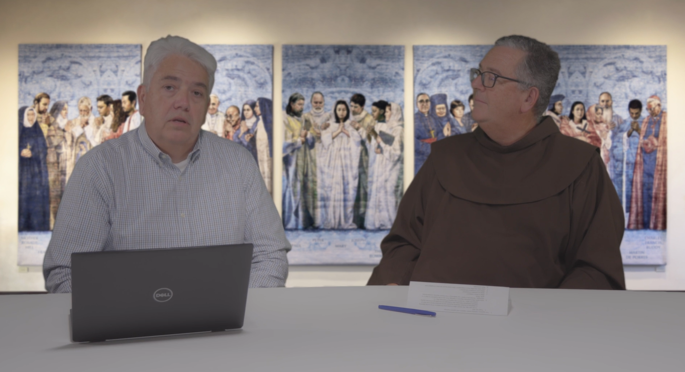 Two men sit at a table with a laptop, facing each other in conversation with artwork displayed.