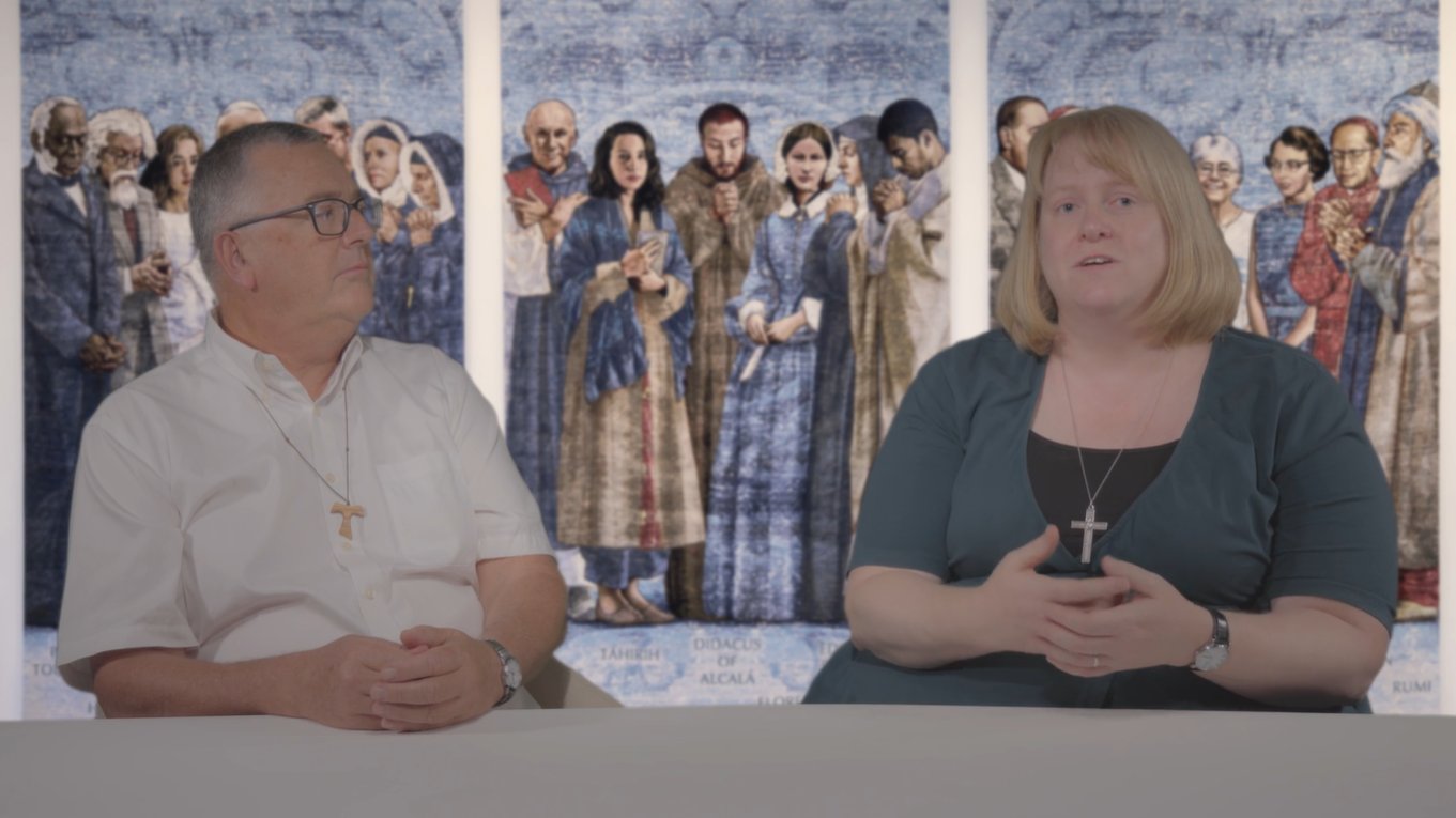 A man and woman sit together in front of a religious mural depicting a group of people.