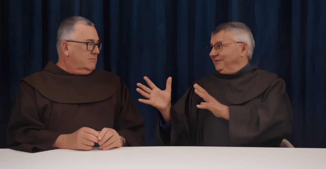 Two men in brown robes wearing glasses sit at a table having a conversation against a blue curtain.