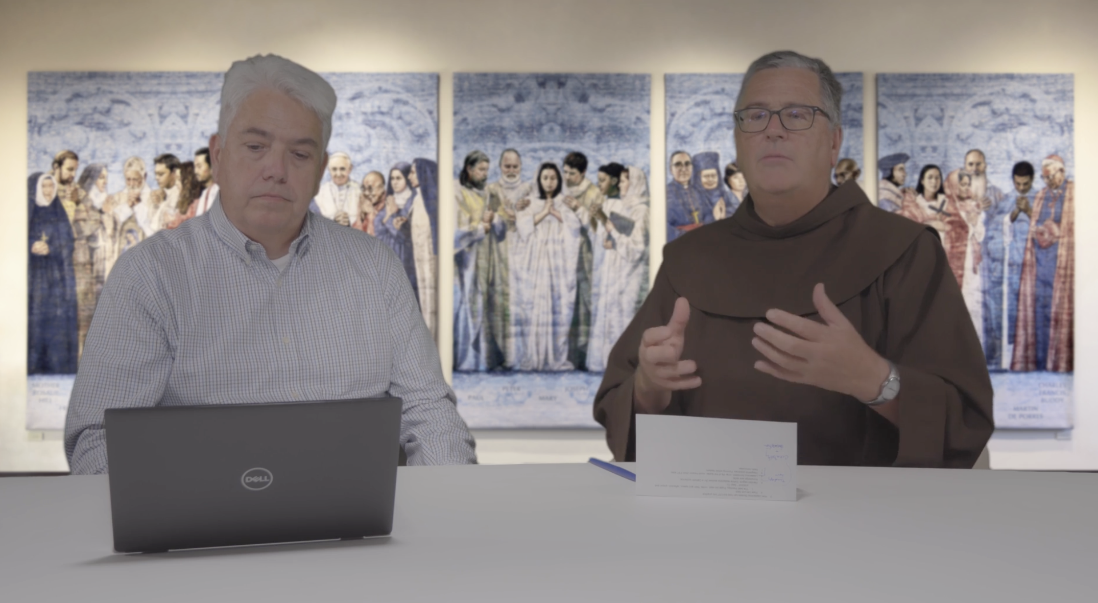 Two men sit at a table with laptops, with religious artwork displayed on the wall behind them.