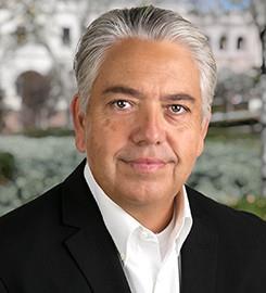 Professional headshot of a middle-aged man with gray hair wearing a dark blazer and white shirt.