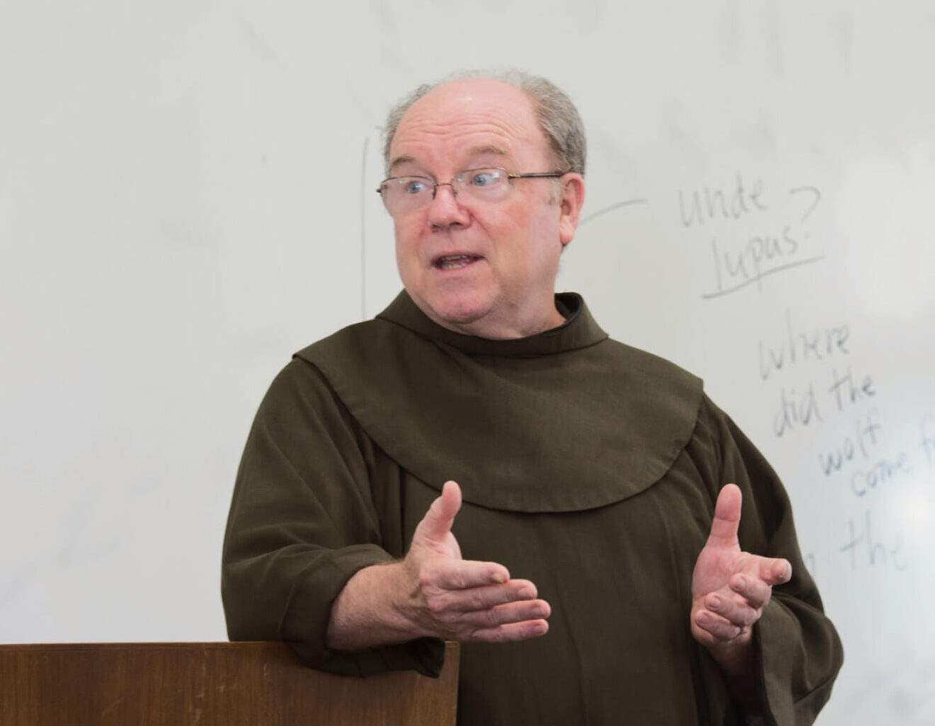 A man wearing glasses and a brown robe speaks at a podium with a whiteboard behind him.