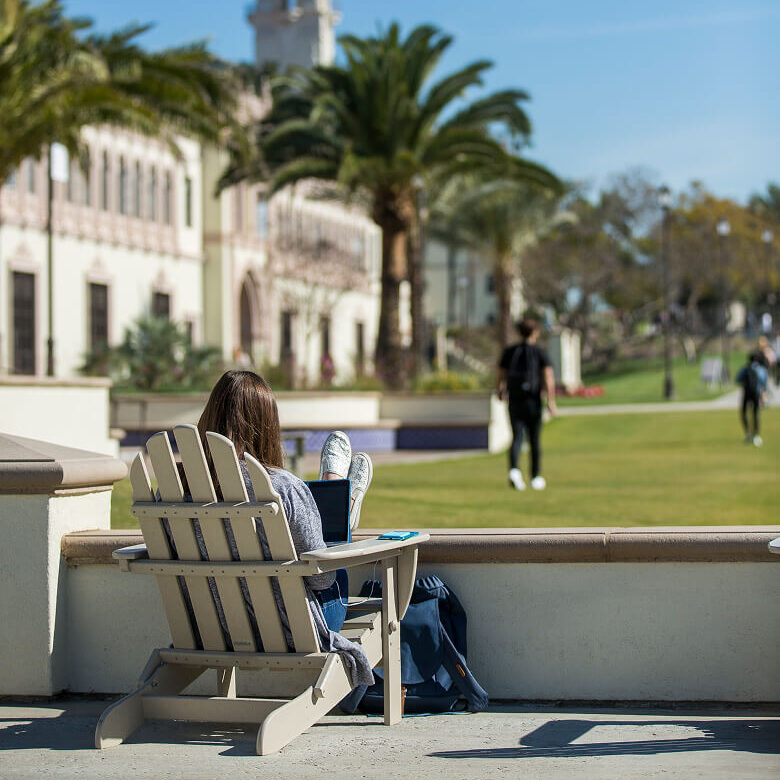 Woman sitting in an Adirondack chair overlooking a campus lawn with palm trees and white buildings.