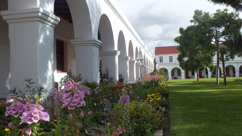 White colonial building with arched colonnade and purple flowers in foreground, green lawn.