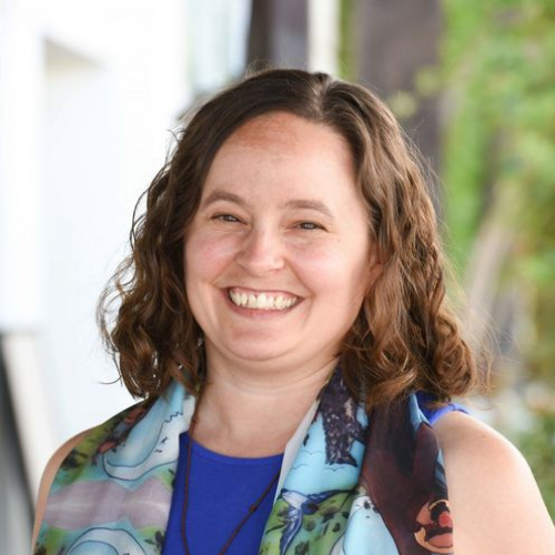 A woman with shoulder-length wavy brown hair smiles at the camera wearing a blue top and patterned.